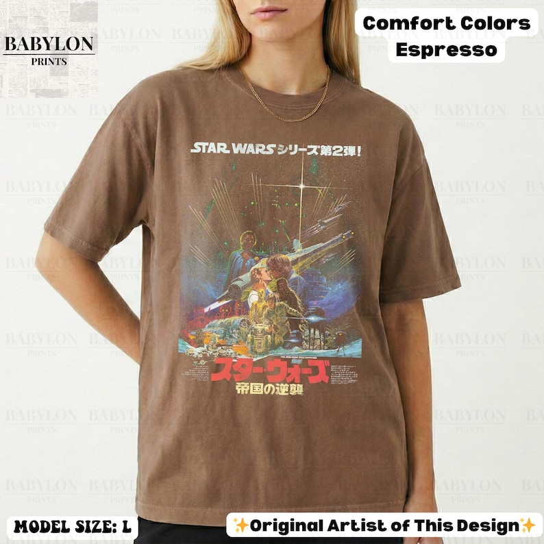 May include: Espresso-colored t-shirt featuring a Star Wars movie poster design. The graphic includes Japanese text and a vintage aesthetic. The shirt is labeled as Comfort Colors and the model is wearing a size L.