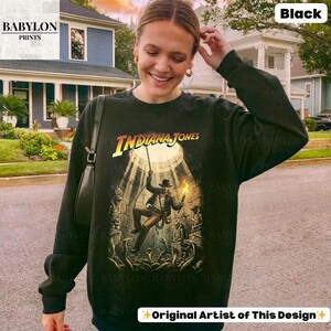 May include: Black sweatshirt featuring the Indiana Jones movie logo and a graphic of the character in a cave. The sweatshirt has long sleeves and a crew neck. The Babylon Prints logo is in the upper left corner.