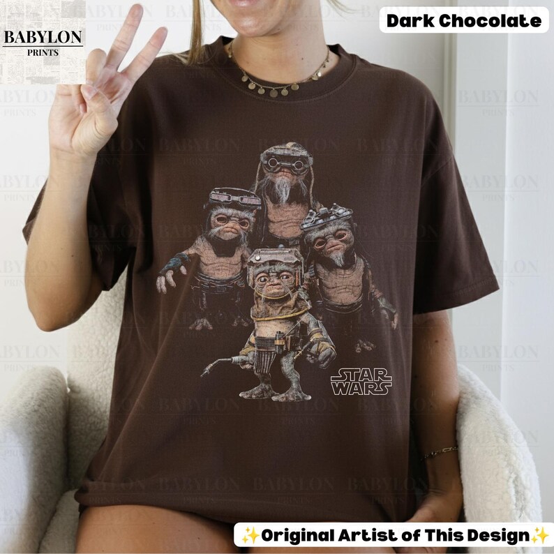 Op de afbeelding: Een donker chocoladebruin t-shirt met korte mouwen en een grafische print van vier Ewok-personages uit Star Wars. De personages zijn afgebeeld in een gedetailleerde, realistische stijl met een bril en gereedschap. Het Star Wars-logo is in het wit gedrukt.