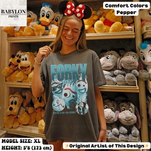 May include: A dark gray t-shirt with the word "FORKY" in light blue, featuring cartoon characters. The shirt is part of the "Comfort Colors" collection. The model is wearing an XL size and is 5'8" (173 cm) tall.