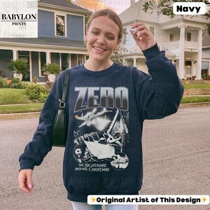 May include: Navy blue sweatshirt featuring the word "ZERO" in a silver and white font, with a graphic from "The Nightmare Before Christmas." The design includes images of Zero, Jack Skellington, and other characters from the movie. The sweatshirt has long sleeves.