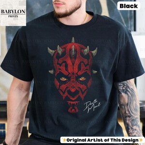 Może przedstawiać: Czarna koszulka z czerwoną grafiką twarzy Darth Maula, z ż&oacute;łtymi oczami i rogami. Tekst "Darth Maul" jest napisany białą czcionką. Koszulka ma u dołu napis "Original Artist of This Design".