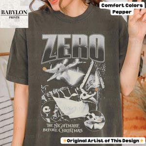 May include: Dark gray t-shirt featuring a graphic from "The Nightmare Before Christmas." The design includes the word "ZERO" in large letters, along with images of Zero and other characters. The text "THE NIGHTMARE BEFORE CHRISTMAS" is also visible.