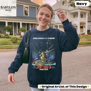 May include: Navy blue sweatshirt featuring a Star Wars movie poster design with Japanese text. The graphic includes images of characters and a spaceship. The sweatshirt has long sleeves and a crew neck. The Babylon Prints logo is in the upper left corner.