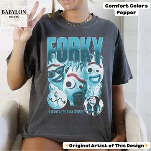 May include: Dark grey t-shirt featuring the word "FORKY" in teal, with cartoon spork characters. The shirt includes the text "I'M NOT A TOY, I'M A SPORK!". The shirt is part of the Comfort Colors collection.