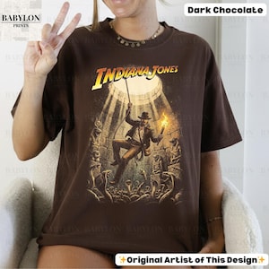 May include: Dark chocolate-colored t-shirt featuring a graphic of Indiana Jones in an adventurous scene. The design includes the text "INDIANA JONES" in a yellow and white font. The image shows a man holding a torch and rope, surrounded by snakes.