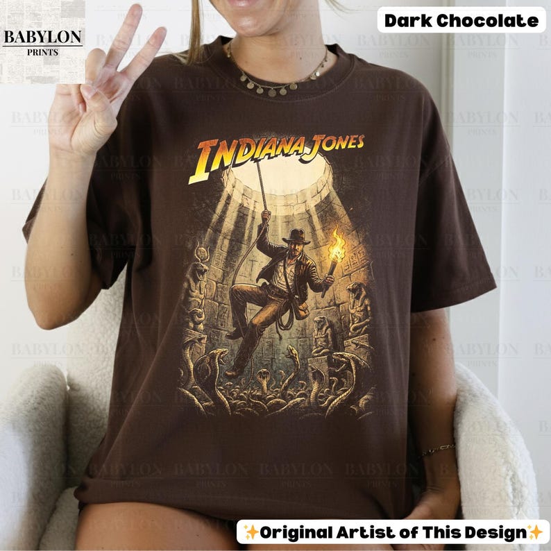 May include: Dark chocolate-colored t-shirt featuring a graphic of Indiana Jones in an adventurous scene. The design includes the text "INDIANA JONES" in a yellow and white font. The image shows a man holding a torch and rope, surrounded by snakes.