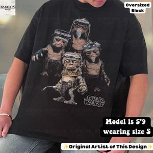 May include: An oversized black t-shirt featuring a graphic print of four Star Wars Ewok characters wearing goggles and gear. The design includes the Star Wars logo in white text at the bottom right. The shirt has short sleeves and a crew neckline.