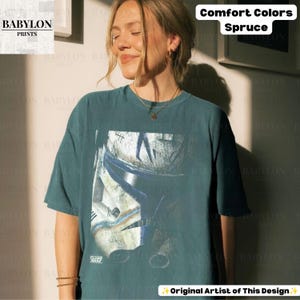 May include: A spruce green short-sleeve t-shirt featuring a graphic print of a weathered white and blue clone trooper helmet. The shirt has a crew neckline and a relaxed fit. The text Comfort Colors Spruce is visible in the upper right corner.