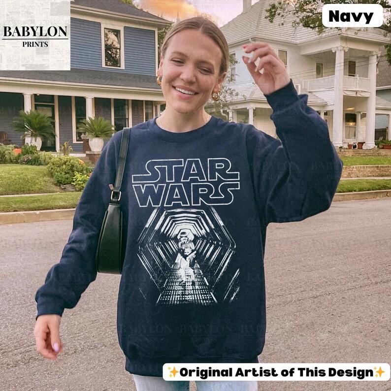 May include: Navy blue Star Wars sweatshirt with a white graphic of the Star Wars logo and a tunnel scene featuring a Stormtrooper. The sweatshirt has long sleeves and a crew neck. The text "Original Artist of This Design" is at the bottom.