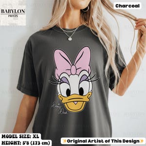 May include: Charcoal-colored t-shirt featuring a large graphic of Daisy Duck with a pink bow, eyelashes, and a yellow beak. The text "Daisy Duck" is written in cursive below the character. The shirt is size XL and the model's height is 5'8 (173 cm).