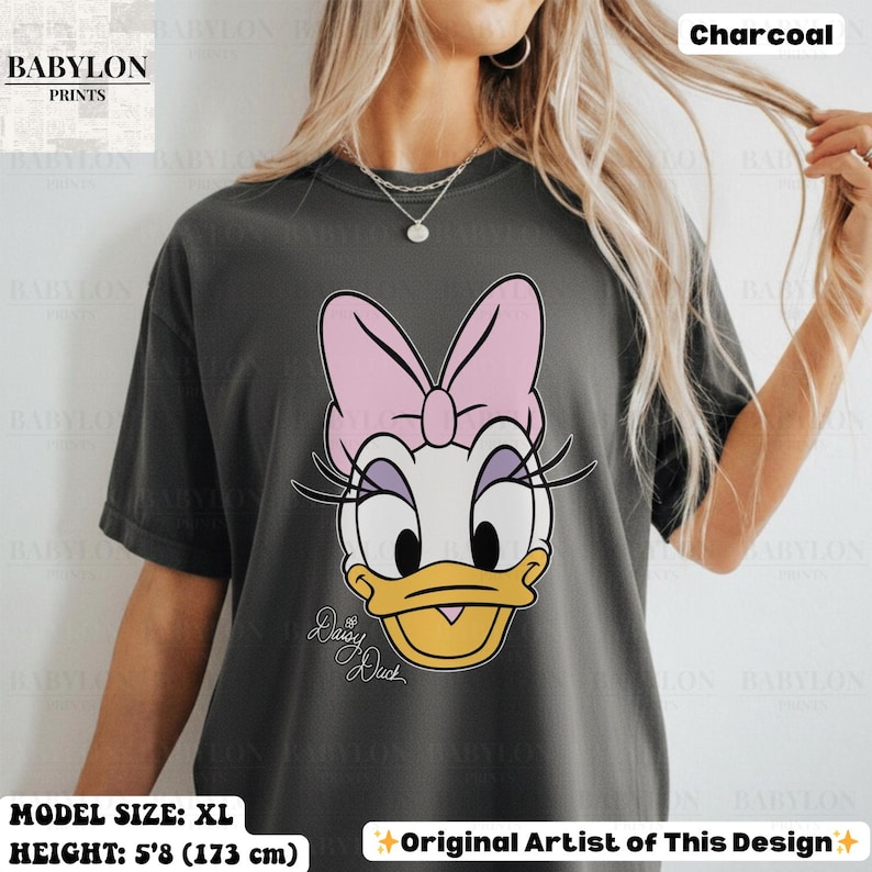 May include: Charcoal-colored t-shirt featuring a large graphic of Daisy Duck with a pink bow, eyelashes, and a yellow beak. The text "Daisy Duck" is written in cursive below the character. The shirt is size XL and the model's height is 5'8 (173 cm).