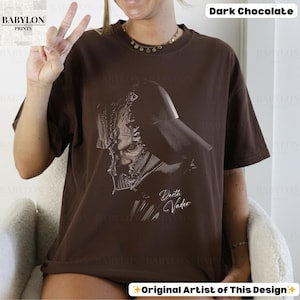 K&ouml;nnte beinhalten: Dunkel schokoladenbraunes T-Shirt mit einer schwarz-wei&szlig;en Illustration von Darth Vader. Der Text "Darth Vader" ist in kursiver Schrift unter dem Bild geschrieben. Das Babylon Prints-Logo ist in der oberen linken Ecke sichtbar.