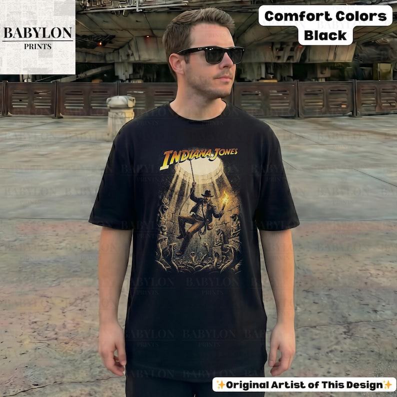 May include: Black t-shirt featuring the Indiana Jones logo and a graphic of the character in a spotlight. The shirt is labeled "Comfort Colors" and "Black" in the upper right corner. The Babylon Prints logo is in the upper left corner.