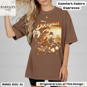 May include: Espresso-colored t-shirt featuring a vintage-style graphic from Disney's DuckTales. The design includes characters and the DuckTales logo. The shirt is a Comfort Colors brand, and the model is wearing a size XL.