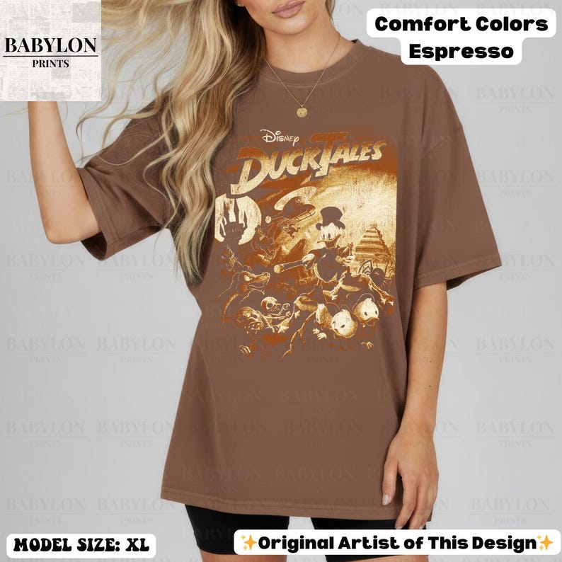 May include: Espresso-colored t-shirt featuring a vintage-style graphic from Disney's DuckTales. The design includes characters and the DuckTales logo. The shirt is a Comfort Colors brand, and the model is wearing a size XL.