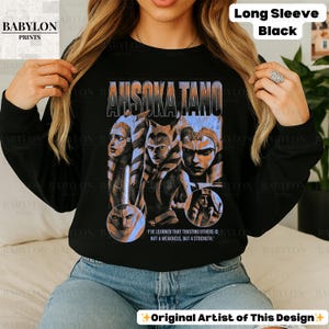 May include: Black long-sleeve sweatshirt with a graphic print of Ahsoka Tano from Star Wars. The graphic is in a vintage style with a blue and white color scheme. The text "I've learned that trusting others is not a weakness, but a strength." is printed below the graphic.