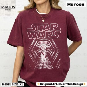 May include: Maroon Star Wars t-shirt with a white graphic. The graphic features the Star Wars logo and a detailed image of characters in a hallway. The shirt is a crew neck and has the text "BABYLON PRINTS" on the top left.