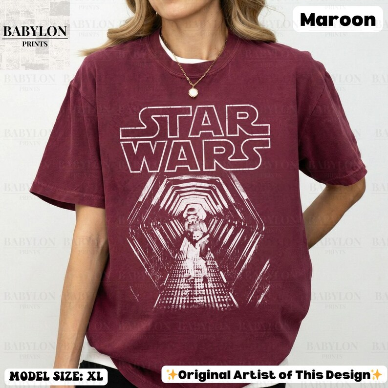 May include: Maroon Star Wars t-shirt with a white graphic. The graphic features the Star Wars logo and a detailed image of characters in a hallway. The shirt is a crew neck and has the text "BABYLON PRINTS" on the top left.