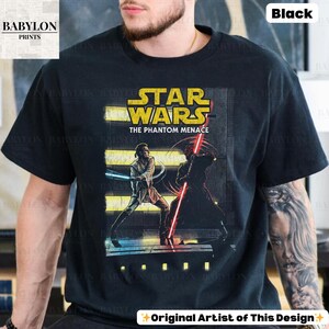 Puede incluir: Camiseta negra con un gr&aacute;fico de Star Wars de "La Amenaza Fantasma". El dise&ntilde;o incluye el logotipo de Star Wars e ilustraciones de personajes con sables de luz. La camiseta tambi&eacute;n tiene el texto "Original Artist of This Design."