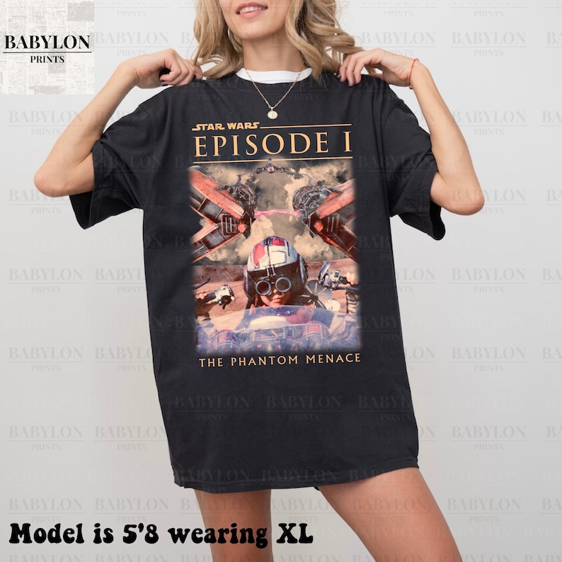 Op de afbeelding: Zwart T-shirt met een Star Wars Episode I-afbeelding. Het ontwerp bevat beelden uit de film, met de tekst "STAR WARS EPISODE I" en "THE PHANTOM MENACE" in goud en wit. Het shirt is donker van kleur.
