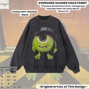 K&ouml;nnte beinhalten: Schwarz gewaschenes Sweatshirt mit einer Grafik von Mike Wazowski aus Monsters, Inc. Das Sweatshirt hat einen Rundhalsausschnitt und lange &Auml;rmel. Der Text "MI" steht auf dem Hut. Das Sweatshirt ist mit "Sweatshirt Washed - Black" gekennzeichnet.