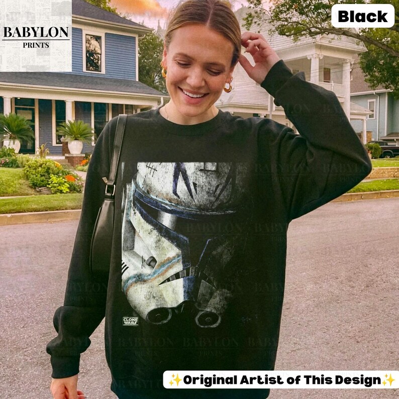 May include: A black long-sleeve sweatshirt featuring a large graphic print of a weathered white and blue clone trooper helmet on the front. The design includes subtle battle-worn details and the text Clone Wars printed near the bottom edge.