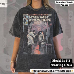 May include: A dark gray t-shirt featuring a comic book cover design from Star Wars: A New Hope. The graphic includes characters and the title. The shirt has a crew neck and short sleeves. The text "BABYLON PRINTS" is visible on the upper left.