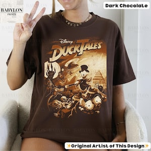 May include: Dark chocolate-colored t-shirt featuring a vintage-style graphic from Disney's DuckTales. The design includes characters like Scrooge McDuck and Huey, Dewey, and Louie, with the text "DuckTales" in a stylized font. The shirt is a relaxed fit.