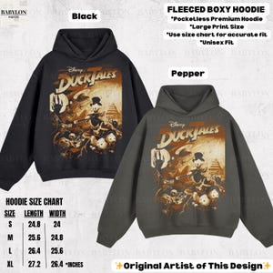 May include: Black and pepper-colored fleece boxy hoodies featuring a Disney DuckTales graphic. The hoodies have a large print size and are unisex fit. The size chart shows measurements in inches. The design is by the original artist.