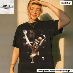 May include: Black t-shirt with a graphic of a woman in a formal gown with her arms raised. The image is a close-up of the woman's face and upper body. The text "BABYLON PRINTS" is in the upper left corner. The text "Black" is in the upper right corner. The text "Original Artist of This Design" is at the bottom.