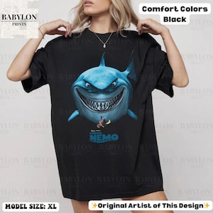 May include: Black t-shirt featuring a large, detailed illustration of a blue shark with a menacing grin, and the text "Finding Nemo". The shirt is from Babylon Prints and is a comfort color.