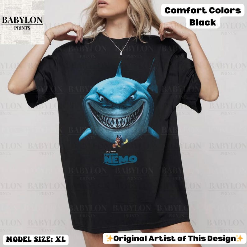 May include: Black t-shirt featuring a large, detailed illustration of a blue shark with a menacing grin, and the text "Finding Nemo". The shirt is from Babylon Prints and is a comfort color.