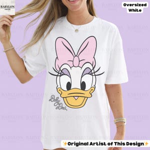 Puede incluir: Camiseta blanca de gran tama&ntilde;o con un dise&ntilde;o de Daisy Duck. El dise&ntilde;o incluye un lazo rosa, pesta&ntilde;as y el texto "Daisy Duck". La camiseta tambi&eacute;n tiene el texto "Original Artist of This Design".