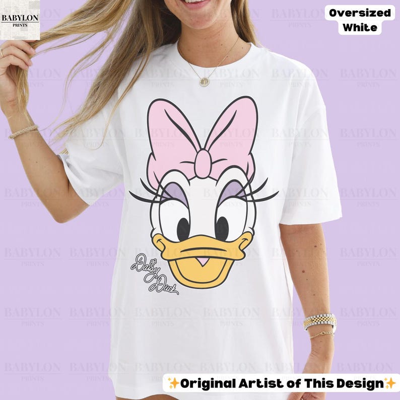 Puede incluir: Camiseta blanca de gran tama&ntilde;o con un dise&ntilde;o de Daisy Duck. El dise&ntilde;o incluye un lazo rosa, pesta&ntilde;as y el texto "Daisy Duck". La camiseta tambi&eacute;n tiene el texto "Original Artist of This Design".