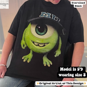 K&ouml;nnte beinhalten: Schwarzes, &uuml;bergro&szlig;es T-Shirt mit einer gro&szlig;en Grafik eines gr&uuml;nen, ein&auml;ugigen Cartoon-Charakters, der einen Hut mit den Buchstaben "MIT" tr&auml;gt. Das Shirt hat den Text "Oversized Black" und "Original Artist of This Design". Das Model ist 1,75 m gro&szlig; und tr&auml;gt Gr&ouml;&szlig;e S.