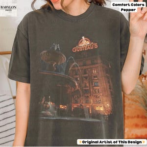 May include: Dark grey t-shirt with a graphic of a fountain and building, and the orange text "GUSTEAU'S". The shirt is labelled "Comfort Colors Pepper" and "Original Artist of This Design".
