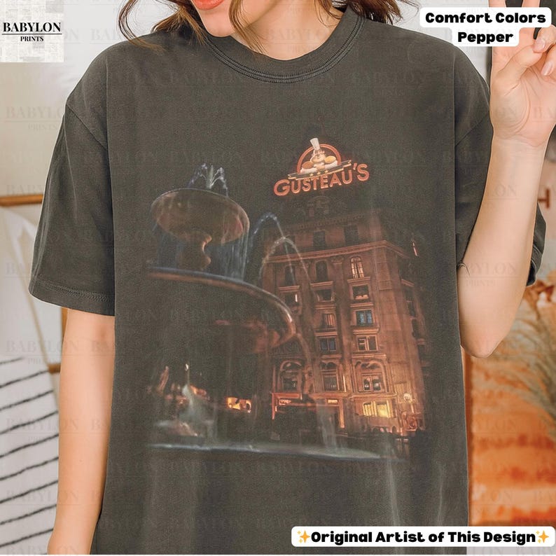 May include: Dark grey t-shirt with a graphic of a fountain and building, and the orange text "GUSTEAU'S". The shirt is labelled "Comfort Colors Pepper" and "Original Artist of This Design".
