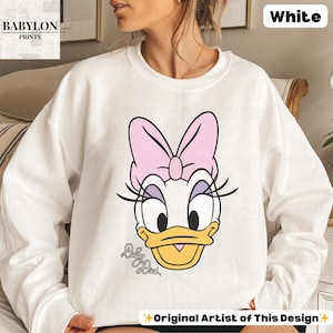 May include: White sweatshirt featuring a Daisy Duck graphic. The design includes Daisy's face with a pink bow, eyelashes, and the text "Daisy Duck". The sweatshirt is from "Babylon Prints" and is labeled "Original Artist of This Design".