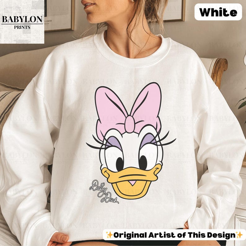 May include: White sweatshirt featuring a Daisy Duck graphic. The design includes Daisy's face with a pink bow, eyelashes, and the text "Daisy Duck". The sweatshirt is from "Babylon Prints" and is labeled "Original Artist of This Design".