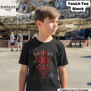 Może przedstawiać: Czarny t-shirt dla młodzieży z czerwoną grafiką twarzy Darth Maula, rogami i napisem "Darth Maul". Koszulka ma kr&oacute;tkie rękawy i okrągły dekolt. Słowa "Youth Tee Black" znajdują się w białym polu w prawym g&oacute;rnym rogu.