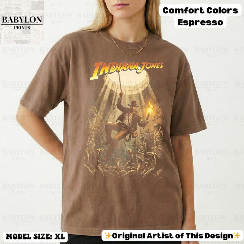 May include: Espresso-colored t-shirt featuring the Indiana Jones movie logo and a graphic of the character in an action scene. The shirt has short sleeves and a relaxed fit. The text "Comfort Colors" and "Original Artist of This Design" are also visible.