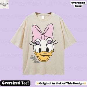 Puede incluir: Camiseta de gran tama&ntilde;o de color arena con un gr&aacute;fico de Daisy Duck. El dise&ntilde;o incluye un lazo rosa, pesta&ntilde;as y el texto "Daisy Duck". La camiseta tiene mangas cortas y cuello redondo. Tambi&eacute;n se ve el texto "Oversized Tee!" y "Original Artist of This Design".