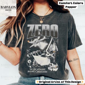 May include: Dark gray t-shirt featuring the word "ZERO" above an illustration of Zero from "The Nightmare Before Christmas." The design includes images of Jack Skellington and other characters from the film. The shirt is labeled "Comfort Colors Pepper."