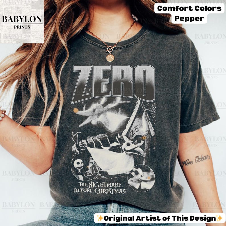 May include: Dark gray t-shirt featuring the word "ZERO" above an illustration of Zero from "The Nightmare Before Christmas." The design includes images of Jack Skellington and other characters from the film. The shirt is labeled "Comfort Colors Pepper."
