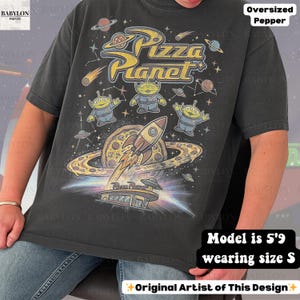 May include: Dark gray oversized t-shirt featuring a Pizza Planet graphic. The design includes the Pizza Planet logo, cartoon aliens, a rocket ship, and a pizza planet. The shirt has a vintage, distressed look.