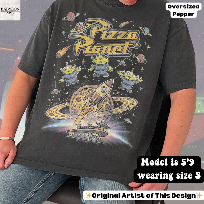 May include: Dark gray oversized t-shirt featuring a Pizza Planet graphic. The design includes the Pizza Planet logo, cartoon aliens, a rocket ship, and a pizza planet. The shirt has a vintage, distressed look.