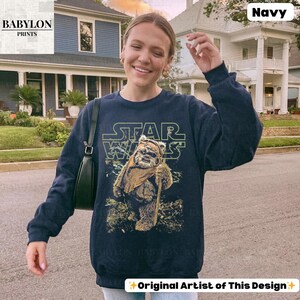 May include: Navy blue sweatshirt featuring a Star Wars design with the text "STAR WARS" in green and a graphic of an Ewok character. The sweatshirt has long sleeves and a crew neck. The words "Original Artist of This Design" are at the bottom.