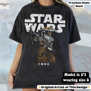 Puede incluir: Camiseta negra de Star Wars con el personaje Embo. La camiseta tiene un aspecto desgastado con texto blanco y una ilustraci&oacute;n detallada de Embo con un arma. La palabra "EMBO" est&aacute; debajo. La modelo mide 1,70 m y usa talla S.