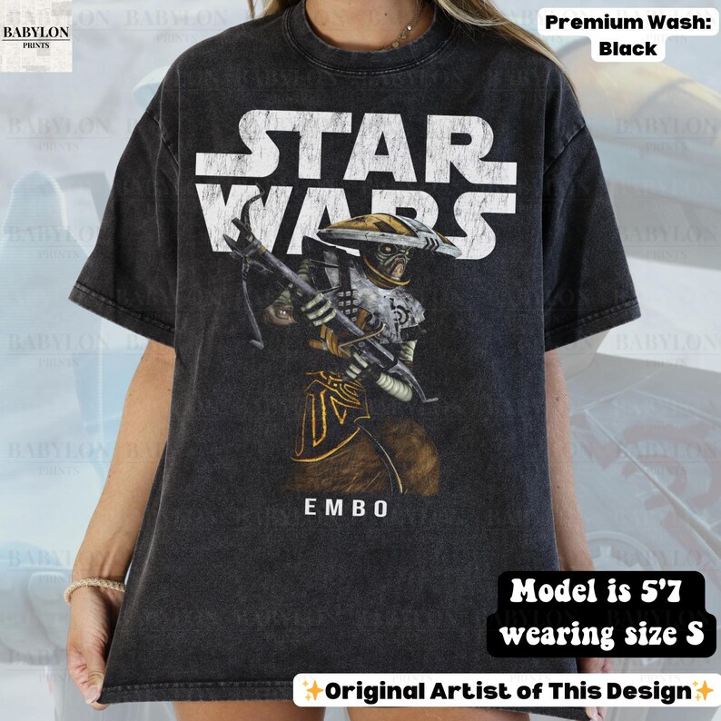 Puede incluir: Camiseta negra de Star Wars con el personaje Embo. La camiseta tiene un aspecto desgastado con texto blanco y una ilustraci&oacute;n detallada de Embo con un arma. La palabra "EMBO" est&aacute; debajo. La modelo mide 1,70 m y usa talla S.